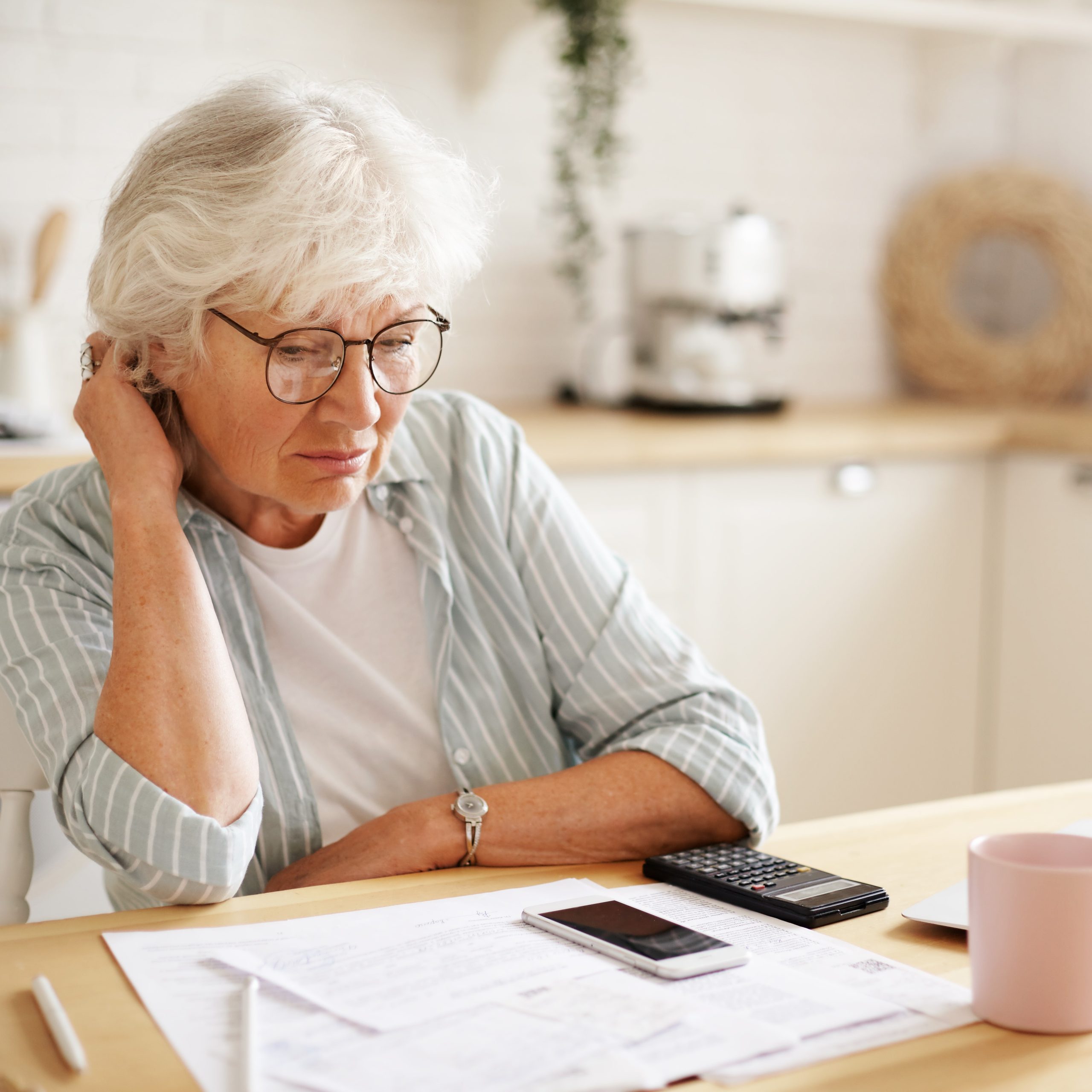 People, age, technology and finances. Depressed unhappy retired woman paying domestic bills online, trying hard to make both ends meet, sitting at kitchen table, surrounded with papers, using gadgets